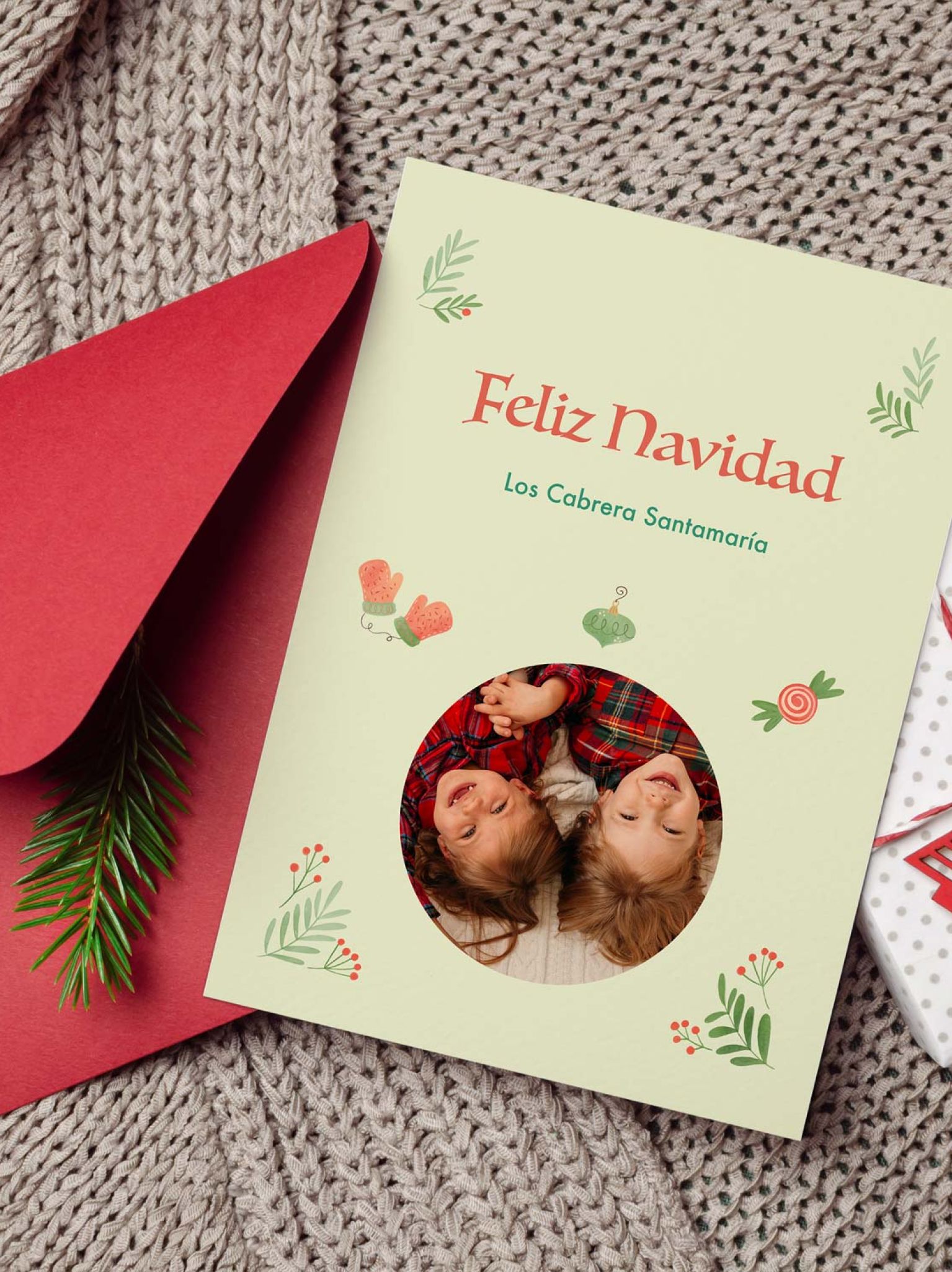 mockup de tarjeta navideña con foto infantil junto a sobre rojo y decoración navideña