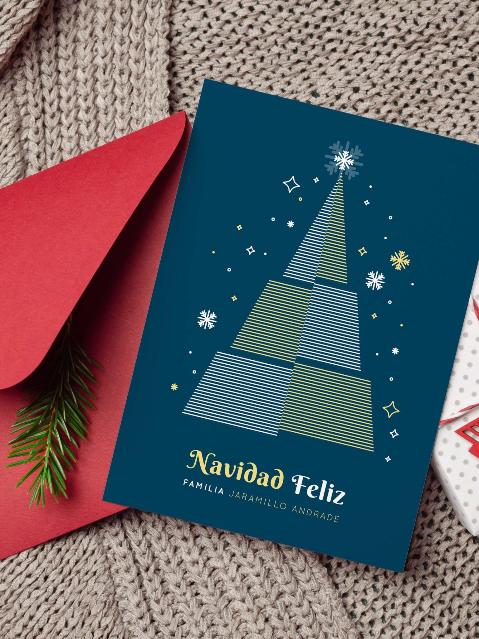 Tarjeta navideña personalizada azul con árbol y sobre rojo, acompañada de decoración navideña y regalo