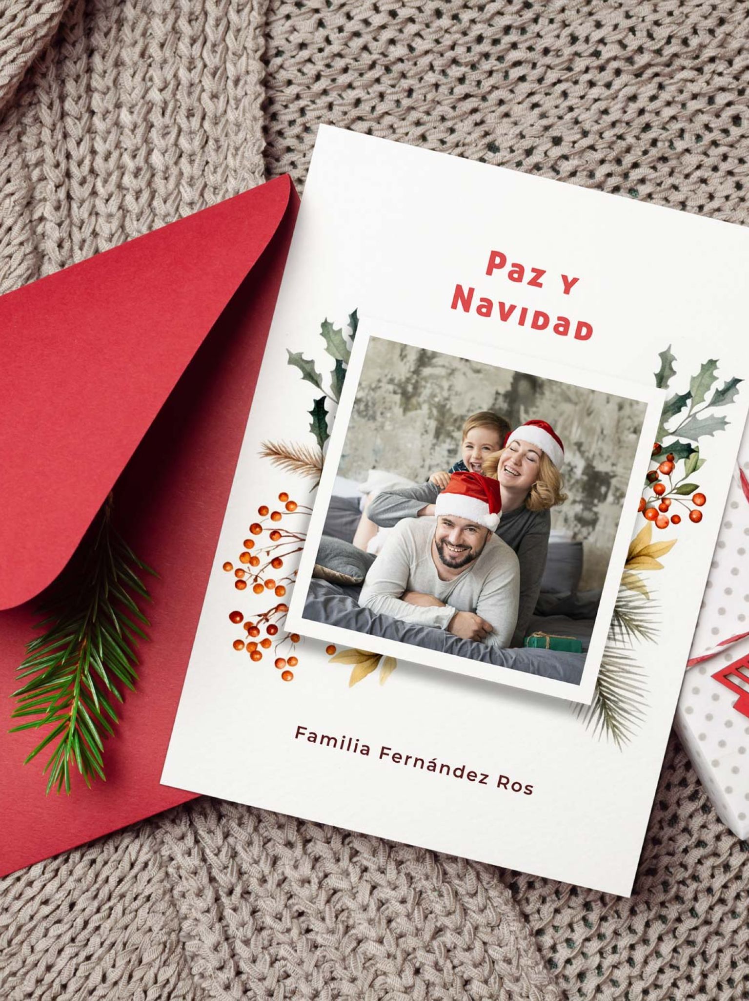 tarjeta navidad con foto personalizada colocada junto a sobre rojo y regalo navide&ntilde;o